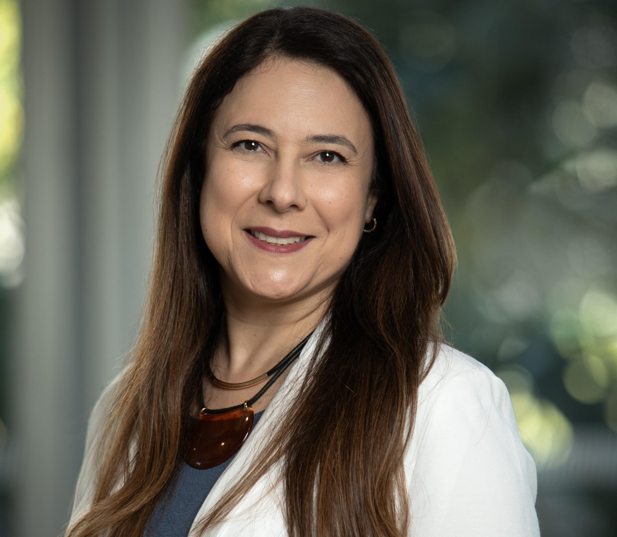  mulher de pele clara, cabelos longos e castanhos, com blusa cinza e blazer branco, sorrindo para a câmera 