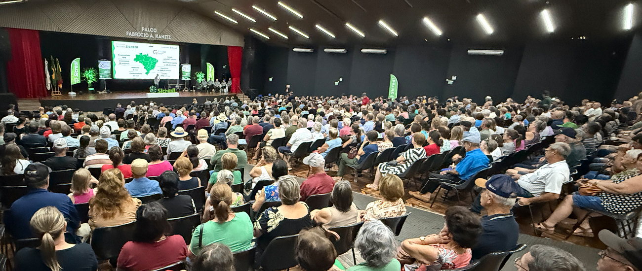  Auditório lotado durante evento promovido pelo Sicredi, com centenas de participantes acompanhando uma apresentação em palco com telão, banners institucionais e conteúdo voltado ao agronegócio e desenvolvimento regional. 