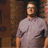  Ricardo está a frente de caixas de produtos, vestindo uma camisa xadrez e de óculos. 