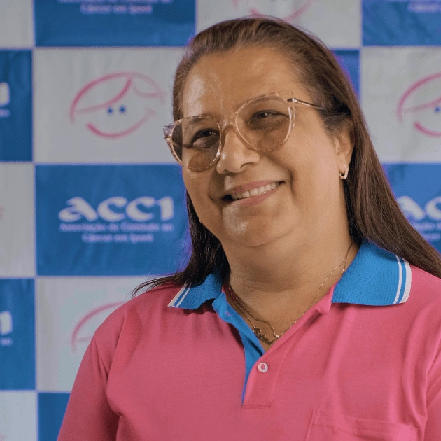  Presidente da ACCI está vestindo uma camiseta rosa e no fundo se encontra um painel com a marca da ONG. 