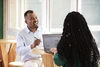  Homem negro conversando com mulher negra, sentados em uma mesa com um computador sobre ela 