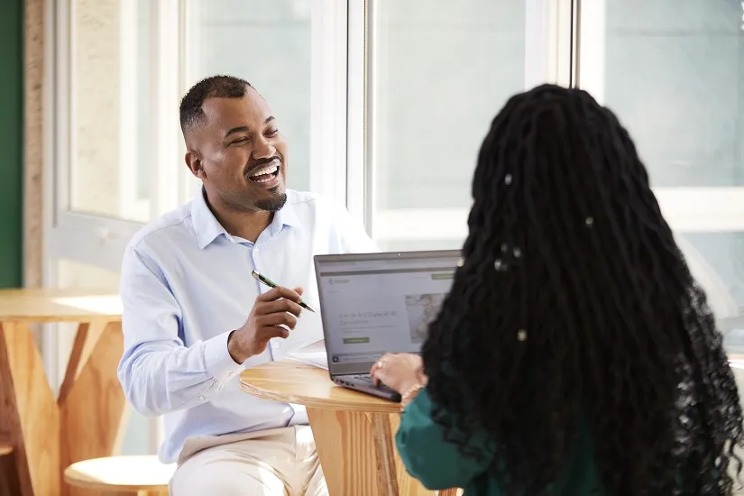  Homem negro conversando com mulher negra, sentados em uma mesa com um computador sobre ela 