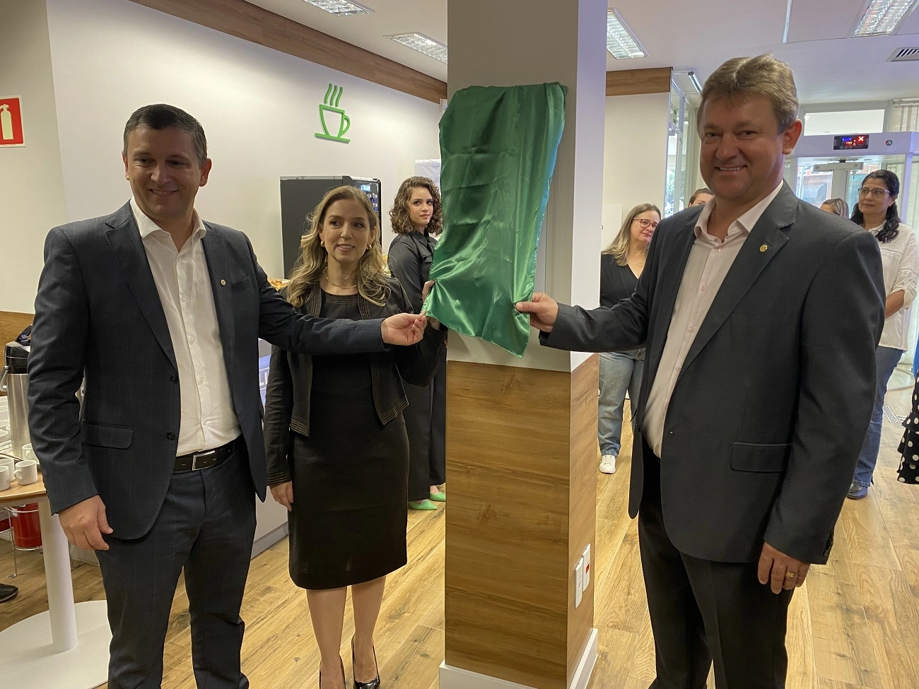  Retirando o tecido da placa de inauguração, 2 homens e 1 mulher com roupas formais. 