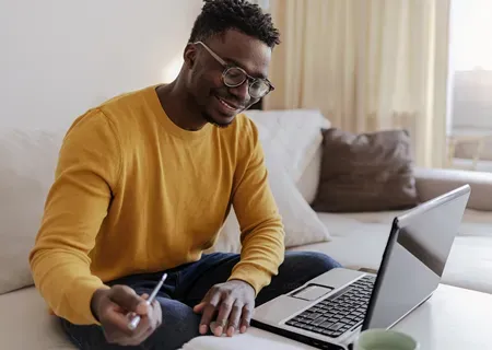  Pessoa sentada em um sofá com um laptop aberto sobre a mesa, segurando uma caneta e um caderno. Há uma caneca ao lado do computador e cortinas claras ao fundo, sugerindo um ambiente doméstico iluminado. 