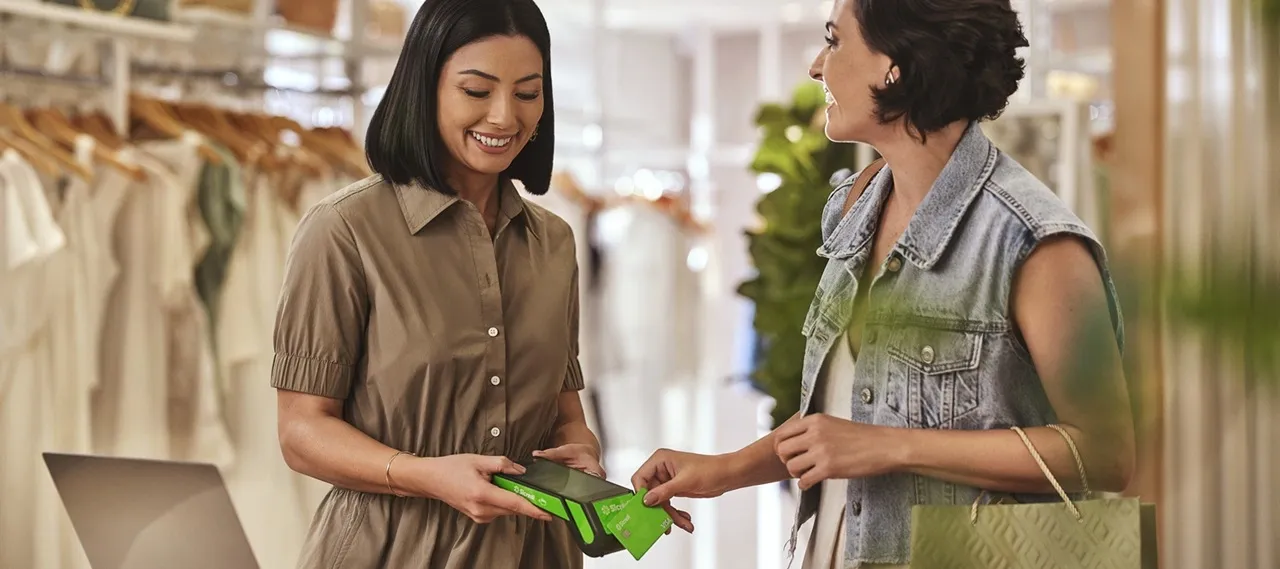  Cena em uma loja de roupas, com duas pessoas interagindo no balcão. Uma delas segura uma maquininha de pagamento verde, enquanto a outra parece realizar uma transação. Ao fundo, há araras com roupas claras e elementos de decoração. 