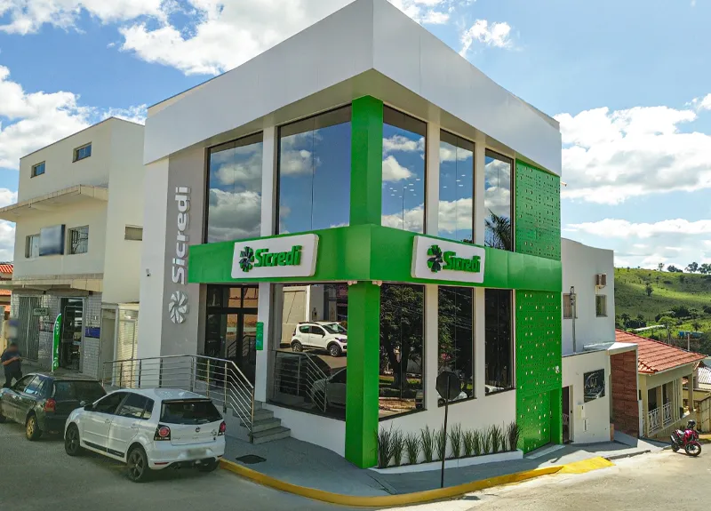  Fachada moderna da agência Sicredi em Matutina/MG, com estrutura em dois andares, grandes painéis de vidro e detalhes verdes característicos da marca, situada em uma rua inclinada em dia ensolarado. 