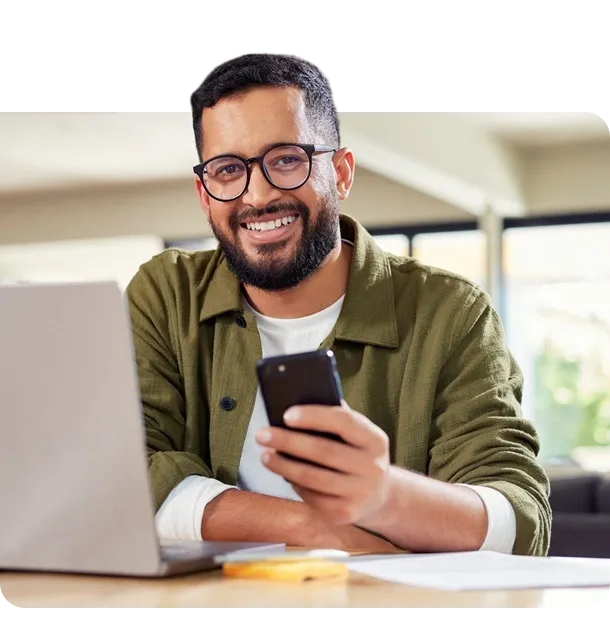  Pessoa sentada à mesa utilizando um notebook enquanto segura um smartphone, em um ambiente interno iluminado. 
