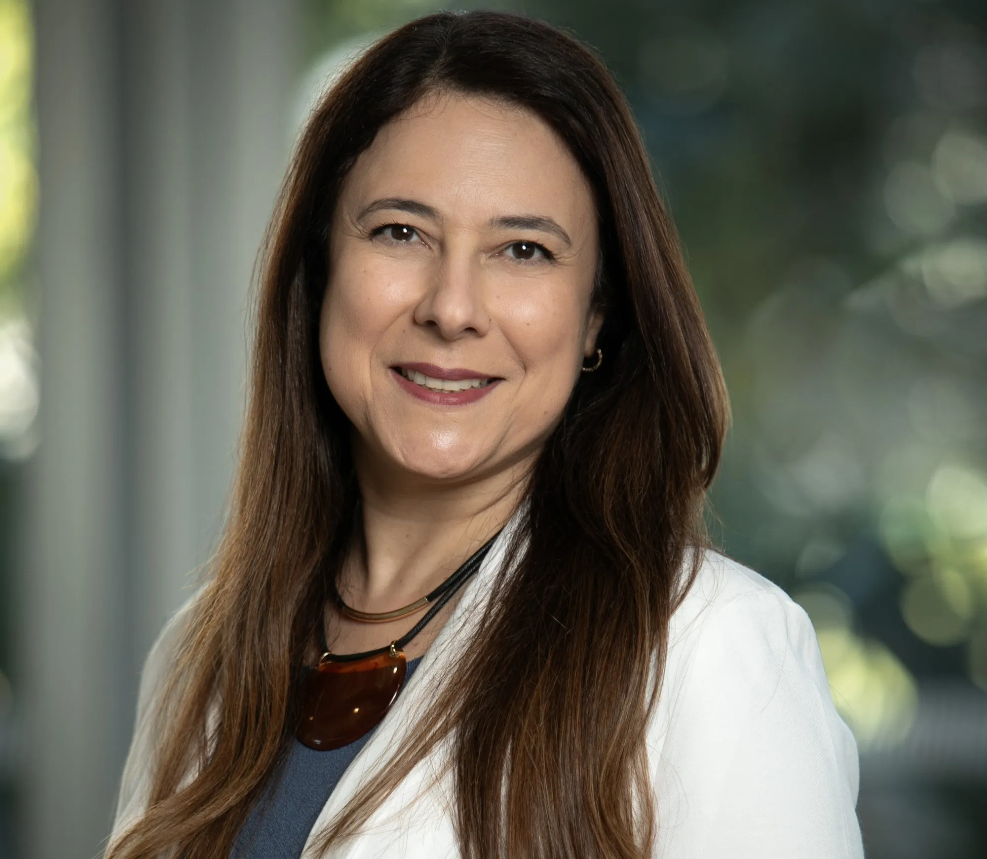  mulher de pele clara, cabelos longos e castanhos, com blusa cinza e blazer branco, sorrindo para a câmera 