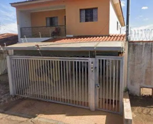  Casa de dois andares com fachada clara, varanda superior, garagem coberta e portão metálico vertical na frente, ao lado de um muro com concertina. A área externa é pavimentada e a garagem está vazia. 