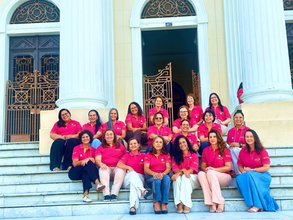  Grupo de mulheres de camisa polo rosa, sentadas em escadaria e sorrindo para a câmera. 