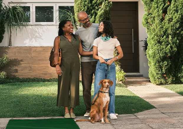  Pessoas adultas em frente a uma casa residencial com jardim, acompanhadas de um cachorro, representando planejamento financeiro, consórcio imobiliário e a conquista da casa própria de forma planejada e sem juros. Qualidade de vida e investimento. 