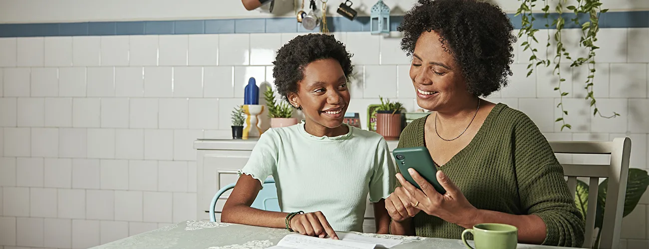  Ambiente interno de uma cozinha ou sala de jantar. Uma pessoa adulta e uma pessoa jovem sentadas à mesa, com um caderno aberto, um telefone celular e uma caneca. A cena remete a orientação, aprendizado e organização financeira no cotidiano familiar. 