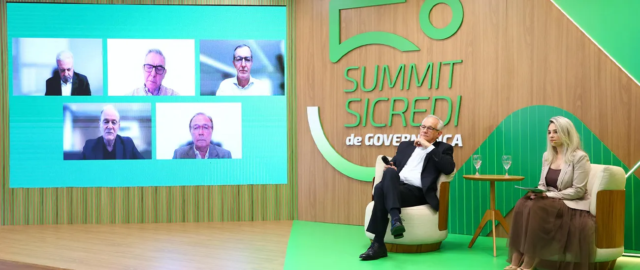  Painel do Summit Sicredi de Governança realizado em formato híbrido, com participantes no palco e convidados conectados por videoconferência em telão, durante debate sobre governança cooperativa. 