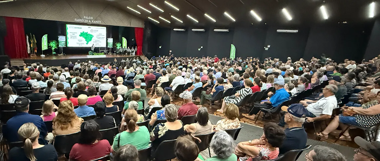  Auditório lotado durante evento promovido pelo Sicredi, com centenas de participantes acompanhando uma apresentação em palco com telão, banners institucionais e conteúdo voltado ao agronegócio e desenvolvimento regional. 