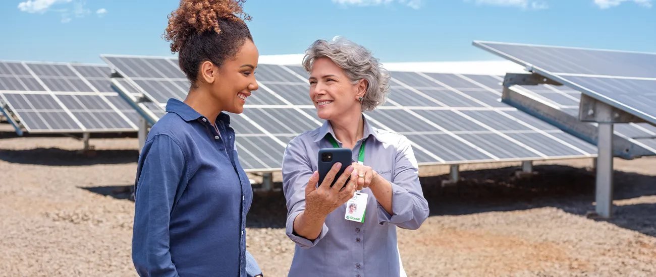 Duas pessoas em área com painéis solares, utilizando dispositivo móvel para acompanhamento de informações, em imagem que representa iniciativas de sustentabilidade, energia renovável e finanças sustentáveis. 