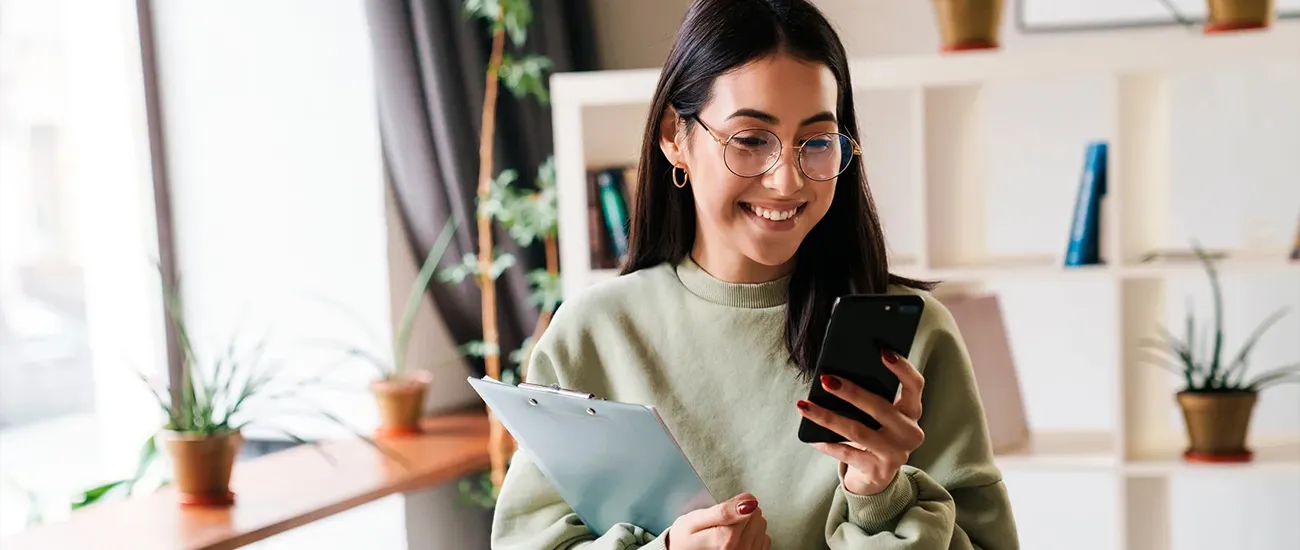  Pessoa utilizando smartphone e segurando uma prancheta em ambiente interno, em imagem utilizada para representar planejamento financeiro, acompanhamento de investimentos e gestão de carteira de investimentos. 