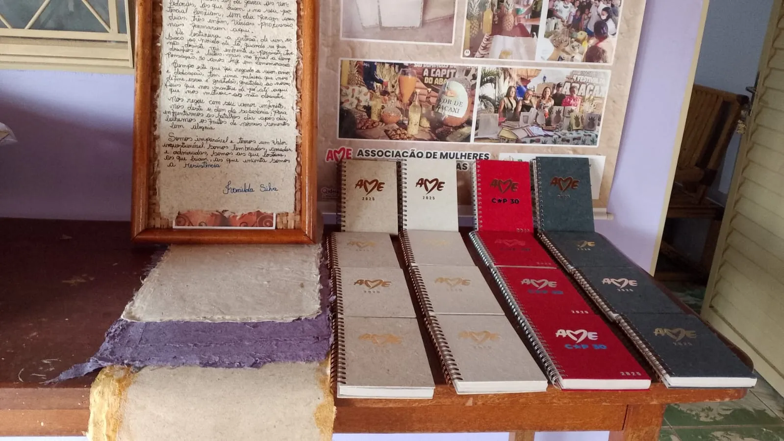  Cadernos, blocos e folhas de papel artesanal produzidos pela AME estão expostos sobre uma mesa, evidenciando o reaproveitamento de materiais e o trabalho desenvolvido pelas artesãs da associação. 