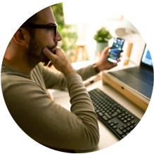  Homem sentado em frente ao computador e com um telefone celular na mão esquerda 