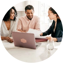  Três pessoas em grupo conversando com papéis em mãos, sentados em uma mesa e em frente a um notebook 