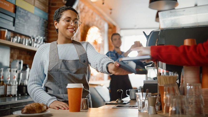  Mulher atrás do balcão e sorrindo, realizando pagamento de cliente através da integração API Pix. 