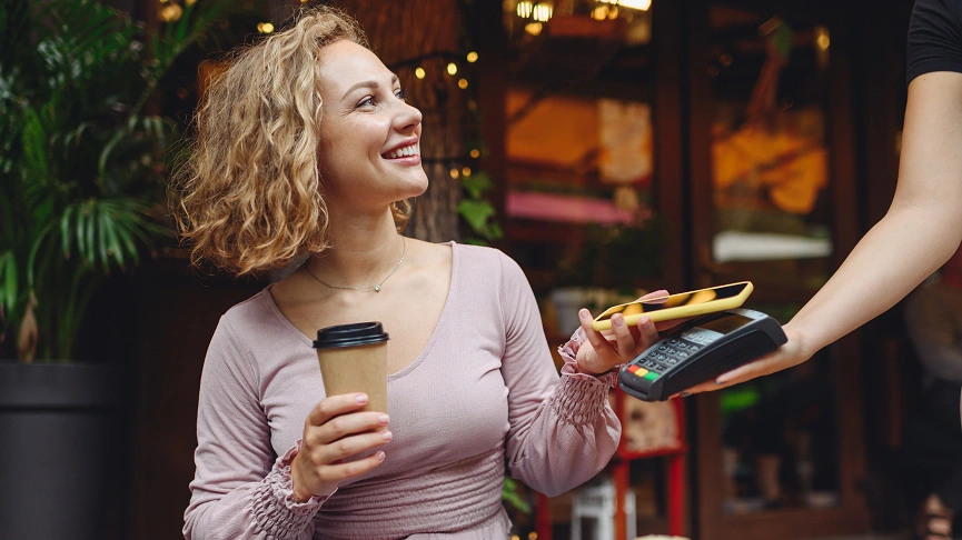  Mulher sentada com um café em mãos, fazendo seu pagamento com carteiras digitais Sicredi. 