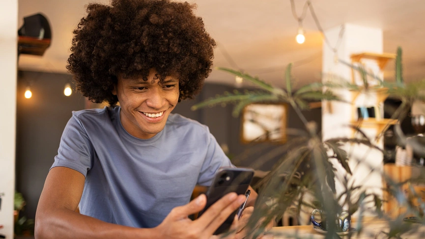  Menino sorrindo e sentado em frente a mesa com celular em mãos, simbolizando a utilização da recarga de celular do Sicredi. 