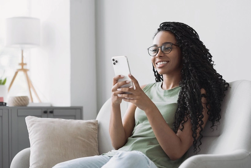 mulher sentada no sofá, usando o celular e sorrindo 