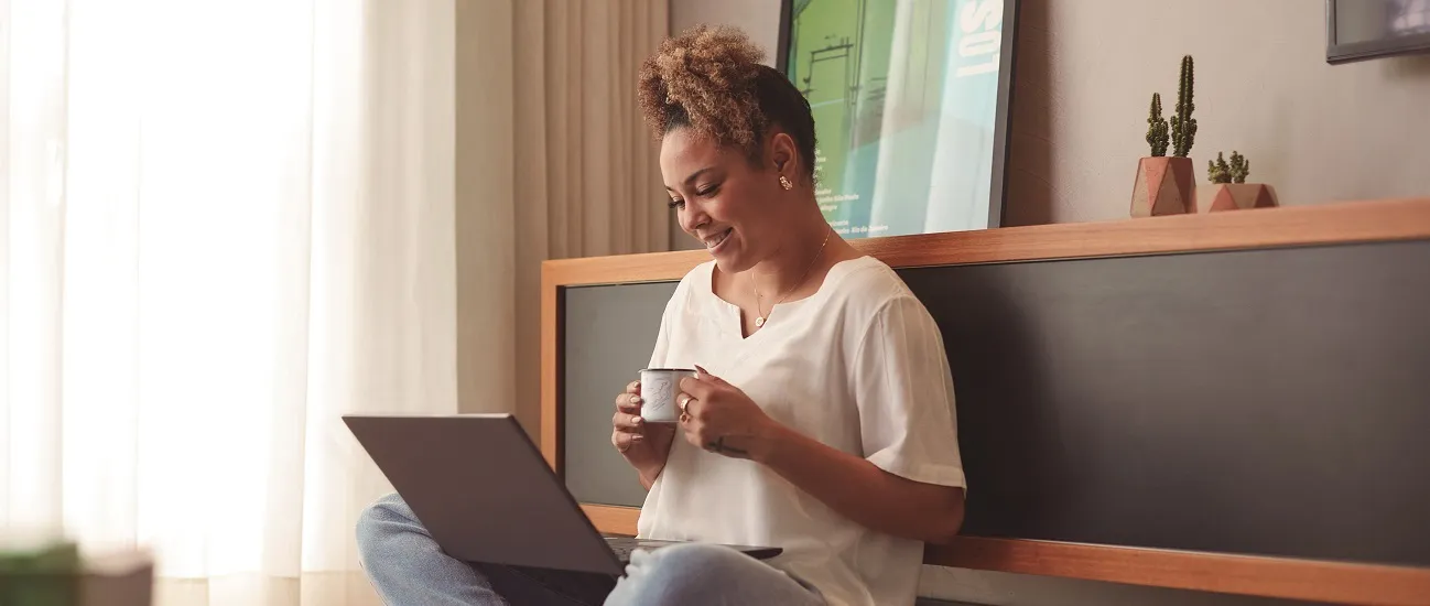  Mulher negra lendo post no Blog do Sicredi enquanto toma uma xicará de café sentada no chão da sua casa. 