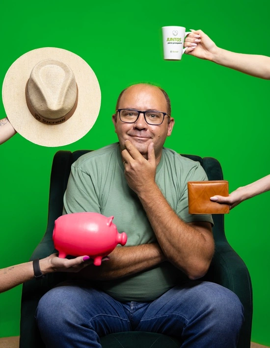  Homem branco careca ao centro, sentado em uma poltrona com produtos de premiação do juntos como canecas e chapéus ao redor dele. 