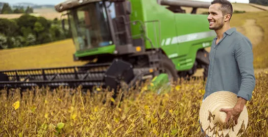 homem segura chapéu de palha em lavoura em frente a um trator