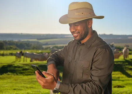 homem negro com chapéu no campo sorri olhando para celular