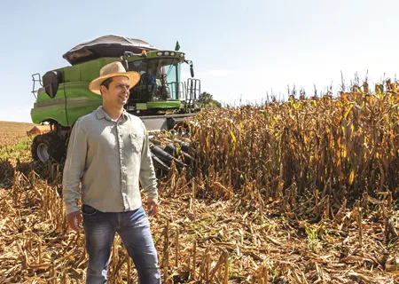 homem branco com chapéu posa em frente a trator e plantação