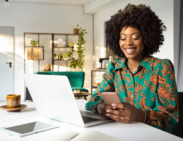  mulher negra sorri enquanto faz compras em notebook utilizando o celular para visualizar seu cartão virtual 
