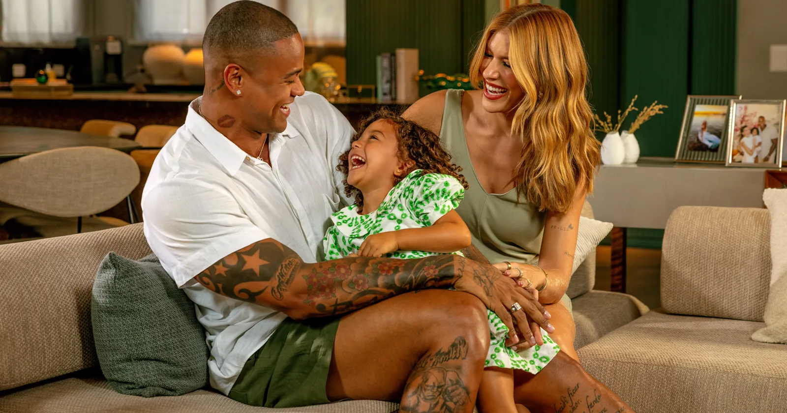  Leo Santana, Lore Improta e filha sorriem em sofá em casa 