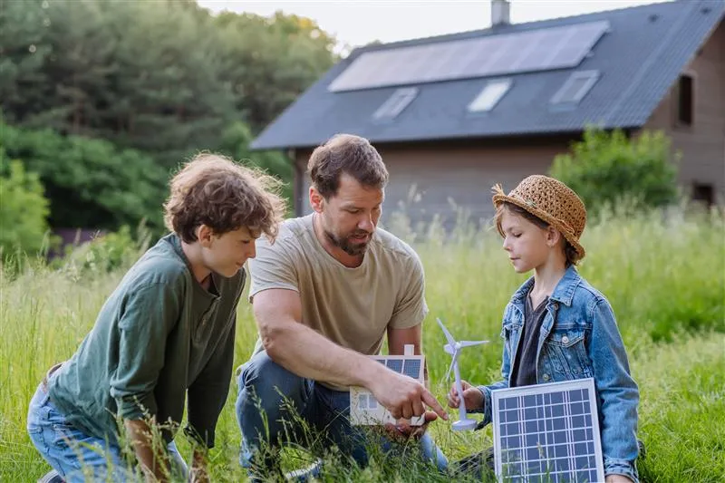  Um pai ensinando seus filhos sobre energia solar no quintal de casa. Ao fundo, a casa com placas solares no telhado. 