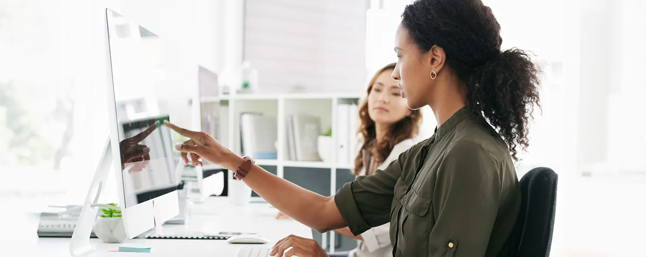  Mulher negra apontando para tela de computador enquanto mulher branca presta atenção 
